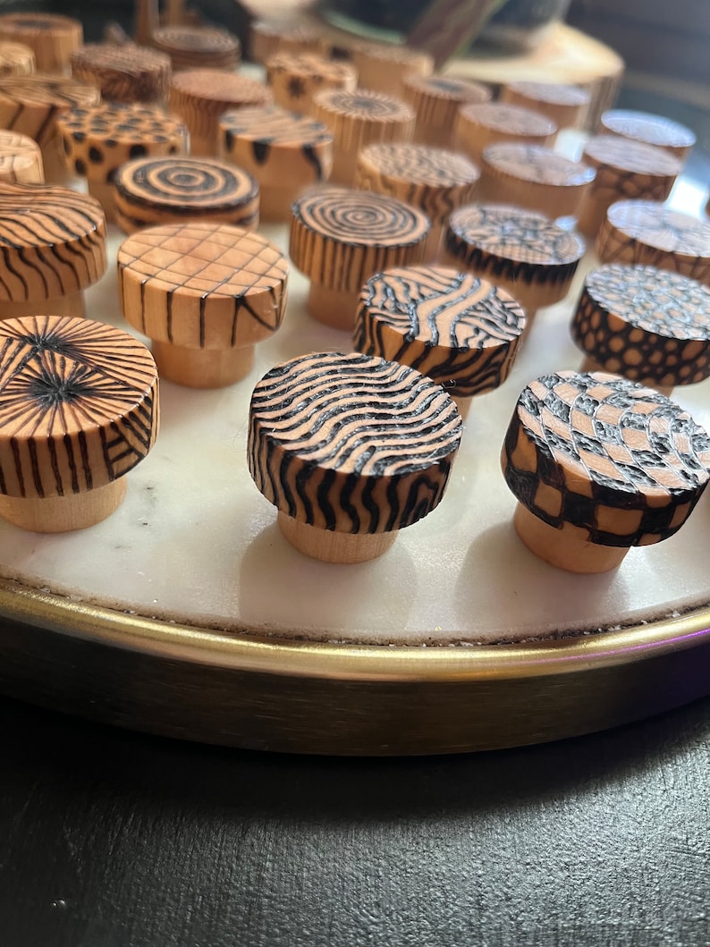 K&ouml;nnte beinhalten: Eine Sammlung von Holzkn&ouml;pfen mit verschiedenen geometrischen Mustern in Schwarz und Wei&szlig;. Die Kn&ouml;pfe sind in einem Kreis auf einer wei&szlig;en Marmorfl&auml;che mit einem goldenen Rand angeordnet.