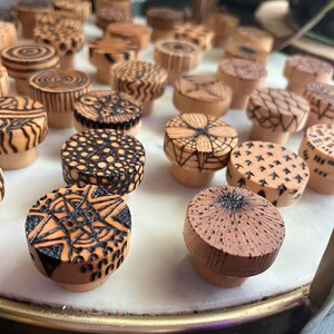 K&ouml;nnte beinhalten: Eine Sammlung von Holzflaschenverschl&uuml;ssen mit komplizierten schwarzen und wei&szlig;en Designs, die in das Holz gebrannt wurden. Die Verschl&uuml;sse sind in einem Kreis auf einer wei&szlig;en Oberfl&auml;che angeordnet.