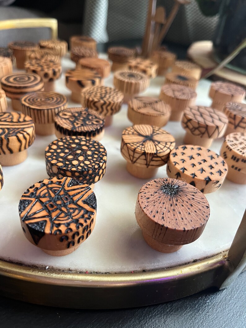 K&ouml;nnte beinhalten: Eine Sammlung von Holzflaschenverschl&uuml;ssen mit komplizierten schwarzen und wei&szlig;en Designs, die in das Holz gebrannt wurden. Die Verschl&uuml;sse sind in einem Kreis auf einer wei&szlig;en Oberfl&auml;che angeordnet.