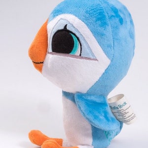 May include: A blue and white plush toy puffin with orange feet and a black eye. The puffin is sitting on a white surface. The toy has a tag that says "Puffin Rock".