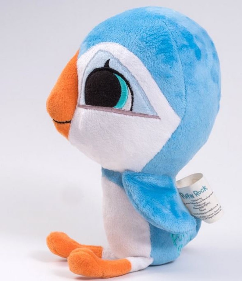 May include: A blue and white plush toy puffin with orange feet and a black eye. The puffin is sitting on a white surface. The toy has a tag that says "Puffin Rock".