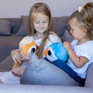 May include: Two young girls are sitting on a couch, each holding a plush toy. The girl on the left is holding a brown and orange plush toy with big eyes. The girl on the right is holding a blue plush toy with a white beak and big eyes.