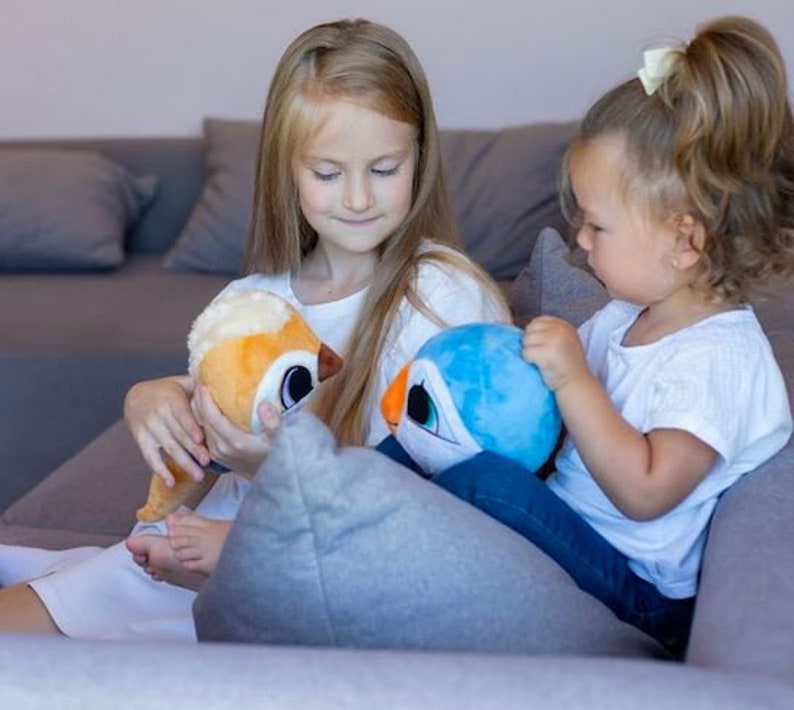 May include: Two young girls are sitting on a couch, each holding a plush toy. The girl on the left is holding a brown and orange plush toy with big eyes. The girl on the right is holding a blue plush toy with a white beak and big eyes.