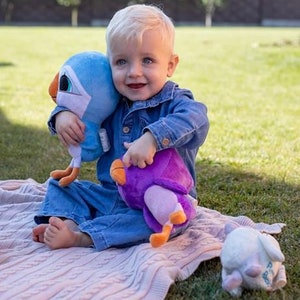 May include: A young child sits on a pink and white blanket holding a blue and white plush bird and a purple plush bird. The child is wearing a blue denim jacket and jeans.
