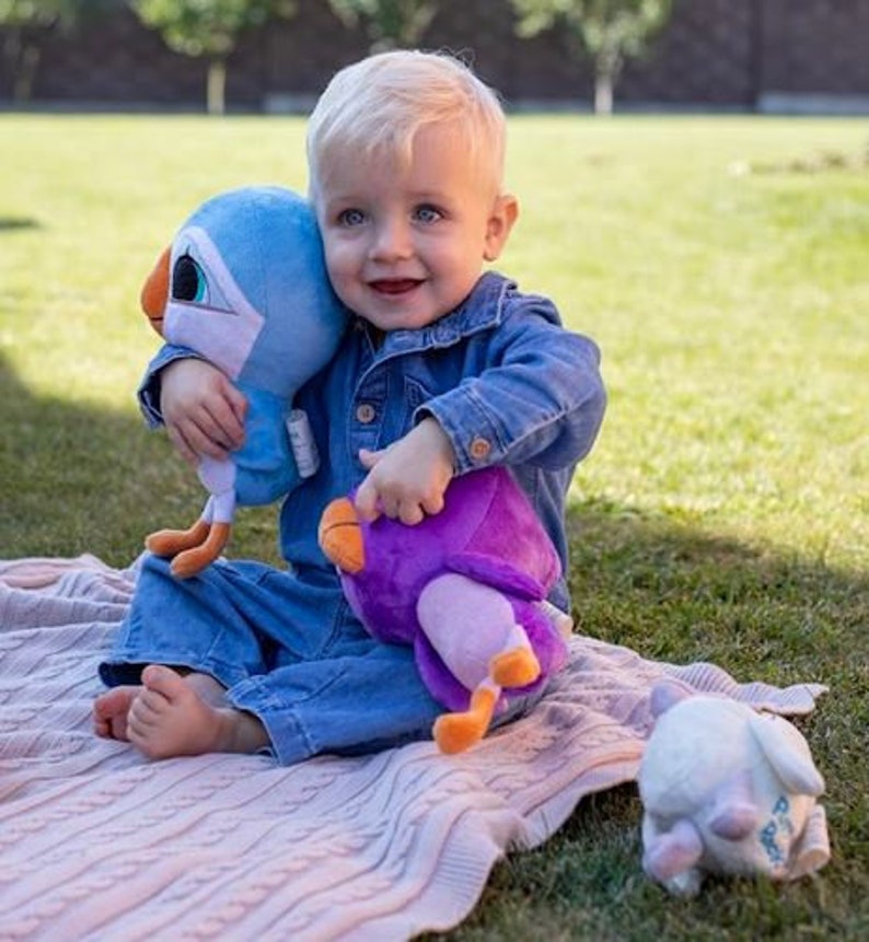 May include: A young child sits on a pink and white blanket holding a blue and white plush bird and a purple plush bird. The child is wearing a blue denim jacket and jeans.