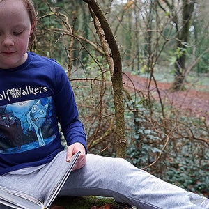 May include: A young person wearing a blue long-sleeved shirt with the words "Wolfwalkers" printed on it, sitting on a tree branch in a forest and reading a book.