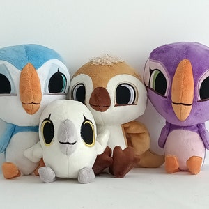 May include: Four plush toys shaped like birds. The birds are all different colors: blue, brown, white, and purple. They have large eyes and beaks. The toys are sitting on a white surface.