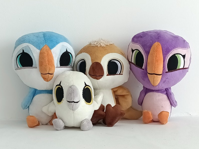 May include: Four plush toys shaped like birds. The birds are all different colors: blue, brown, white, and purple. They have large eyes and beaks. The toys are sitting on a white surface.