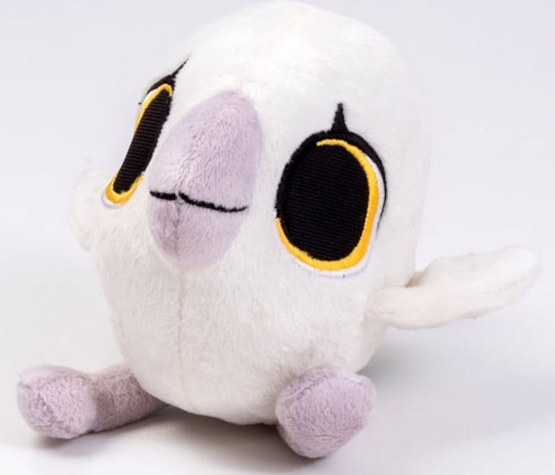 May include: White plush toy with large black eyes, yellow eyelids, and a purple beak. The toy has small purple feet and a white wing.