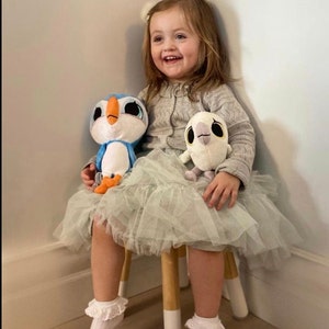 May include: A young girl wearing a light gray sweater and a green tulle skirt sits on a wooden stool. She is holding two plush toys, a blue and white penguin and a white owl. The girl is smiling.