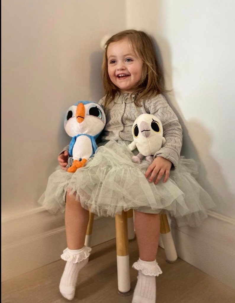 May include: A young girl wearing a light gray sweater and a green tulle skirt sits on a wooden stool. She is holding two plush toys, a blue and white penguin and a white owl. The girl is smiling.