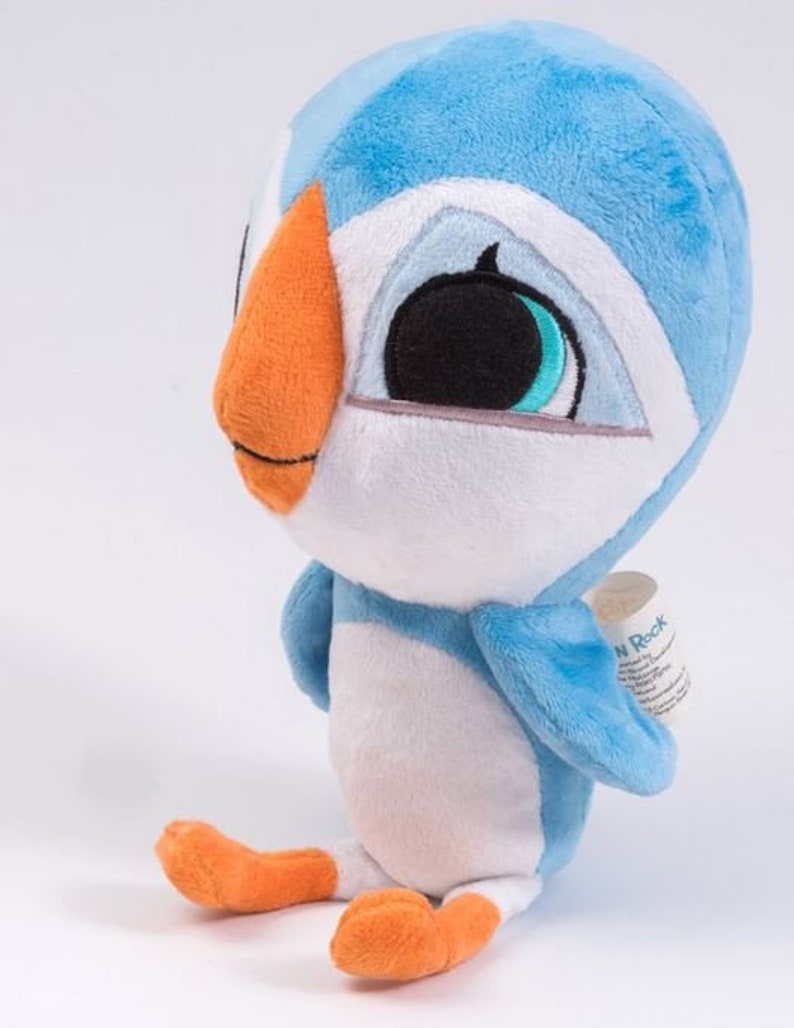 May include: A blue and white plush toy shaped like a puffin with orange feet and beak. The toy has large black eyes with blue accents and a white face. The toy is sitting on a white surface.