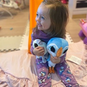 May include: A young girl wearing purple pajamas with a penguin print is sitting on a pink ruffled rug. She is holding two plush toys, one blue and one white.