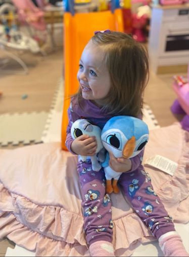 May include: A young girl wearing purple pajamas with a penguin print is sitting on a pink ruffled rug. She is holding two plush toys, one blue and one white.