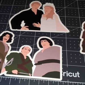 May include: Four illustrated stickers featuring characters from the movie 'The Princess Bride'. The stickers are on a black and white cutting mat. The stickers are white with a white outline. The first sticker shows a man and a woman in wedding attire. The second sticker shows a man with long dark hair and a brown jacket. The third sticker shows a man with a green shirt and a sword. The fourth sticker shows a man with a brown shirt and a belt.