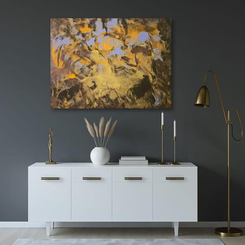 Puede incluir: Una pintura abstracta con tonos dorados, marrones y azules cuelga sobre un mueble blanco. El mueble tiene tiradores dorados y est&aacute; decorado con un jarr&oacute;n blanco, hierba de la pampa y candelabros dorados. Una l&aacute;mpara de pie dorada est&aacute; a la derecha.