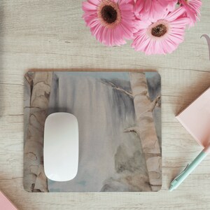 May include: A white computer mouse sits on a mousepad with a watercolor painting of a waterfall and trees. The mousepad is on a light wood surface with a pink notepad and pen, a laptop, and pink flowers.