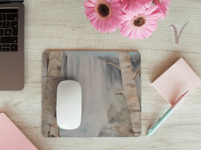 May include: A white computer mouse sits on a mousepad with a watercolor painting of a waterfall and trees. The mousepad is on a light wood surface with a pink notepad and pen, a laptop, and pink flowers.