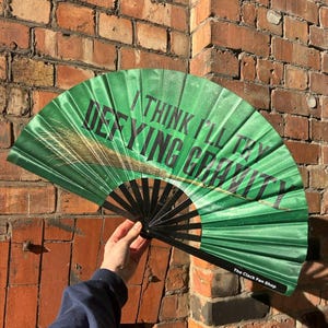 以下が含まれることがあります： 黒いリブが付いた緑色の扇子で、黒で「I THINK I'LL TRY DEFYING GRAVITY」というテキストが特徴です。扇子には金と茶色のイラストと、白で「The Clack Fan Shop」というテキストがあります。