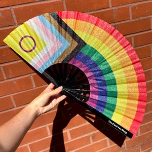 Puede incluir: Un abanico de mano con un diseño de bandera del orgullo intersexual inclusivo con purpurina en un lado y un patrón de arcoíris vibrante en el otro. El abanico tiene varillas negras y el texto The Clack Fan Shop impreso en blanco en el borde.
