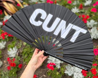 May include: A black hand fan featuring the word CUNT in large white capital letters across the top. The fan has a black frame and includes the text You Betta Merch printed in small white letters on the lower right side of the fabric material.