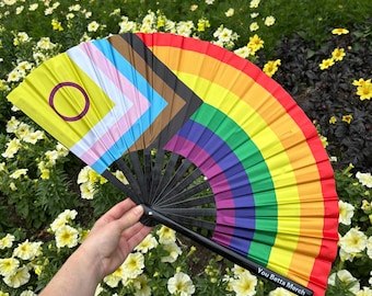 May include: A folding hand fan featuring a split design with the Progress Pride flag on one side and a classic rainbow flag on the other. The fan has black plastic ribs and the text You Betta Merch printed in white on the bottom frame.