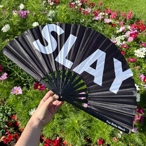 May include: A black folding hand fan featuring the word SLAY in large white capital letters across the fabric. The fan has black ribs and a small label on the bottom right corner that reads You Betta Merch in white text on a black background.
