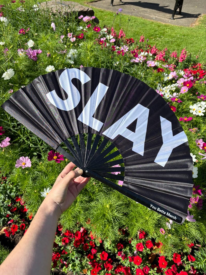 May include: A black folding hand fan featuring the word SLAY in large white capital letters across the fabric. The fan has black ribs and a small label on the bottom right corner that reads You Betta Merch in white text on a black background.