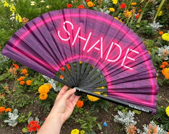 May include: A folding hand fan with a black frame and a dark purple fabric surface featuring a bright pink neon-style arch and the white text SHADE. The text You Betta Merch is printed in small white letters on the lower right side of the frame.