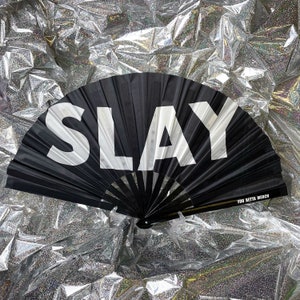 May include: A black folding hand fan featuring the word SLAY in large white capital letters across the fabric. The fan has black ribs and includes the text YOU BETTA MERCH printed in small white letters on the lower right side of the frame.