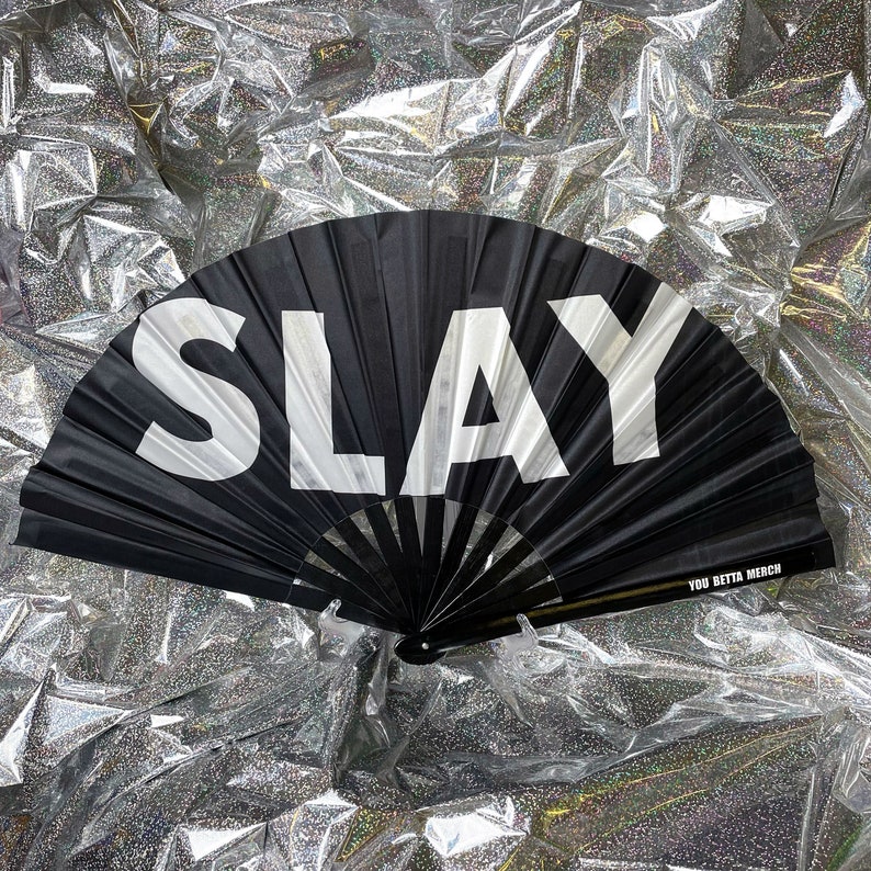 May include: A black folding hand fan featuring the word SLAY in large white capital letters across the fabric. The fan has black ribs and includes the text YOU BETTA MERCH printed in small white letters on the lower right side of the frame.
