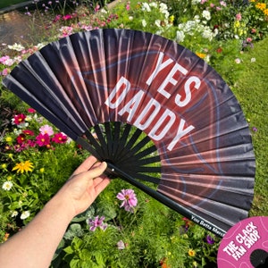 Pode incluir: Um leque dobrável preto e vermelho com o texto "YES DADDY" impresso em branco. O leque está aberto e sendo segurado por uma mão. O fundo é uma imagem desfocada de um jardim de flores.