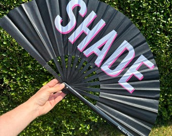 May include: A black hand fan featuring the word SHADE printed in large white capital letters with a bright pink outline. The fan has a black frame and includes the text The clack Fan Shop printed in small white letters on the bottom right corner.