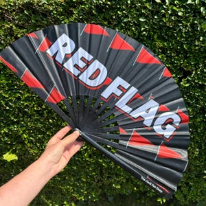 May include: A black hand fan featuring the text RED FLAG in large white capital letters with a red outline. The fan is decorated with multiple small red triangular flag icons scattered across the black background and includes the text The clack Fan Shop.