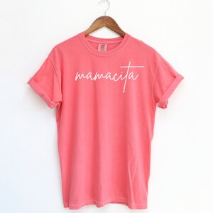 May include: A coral pink t-shirt with the word "mamacita" written in white cursive script.
