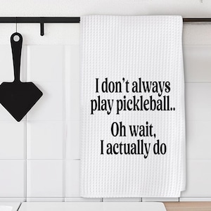 May include: A white waffle-weave kitchen towel featuring the black printed text: I don't always play pickleball.. Oh wait, I actually do. The rectangular towel hangs from a black metal rod mounted on a white tiled wall next to a black utensil.