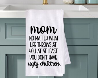 May include: White waffle-weave kitchen towel with the text "mom NO MATTER WHAT LIFE THROWS AT YOU, AT AT LEAST YOU DON'T HAVE ugly children." in black. The towel hangs from a gold faucet in a kitchen setting.