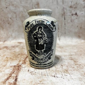 May include: A vintage-style off-white ceramic cream jar featuring a black printed label. The label displays the text Pure Fresh Cream from the Wigtownshire Creamery Co Stranraer, with an illustration of a person holding a bucket in the center.