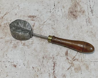 May include: A metal leaf-shaped tool with a wooden handle. The leaf imprint is detailed with veins. The handle is dark brown with a brass fitting. The tool appears to be used for crafting or decorative purposes.