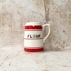 May include: A vintage white ceramic flour canister with red stripes and the word "FLOUR" in black lettering. The canister has a handle and a slightly rounded shape, evoking a classic kitchen aesthetic.