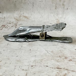 May include: A vintage silver-toned letter opener with a detailed hand and wing design. The opener features a blade and a clip mechanism, likely for holding papers. The design is ornate and the metal has a slightly aged appearance.