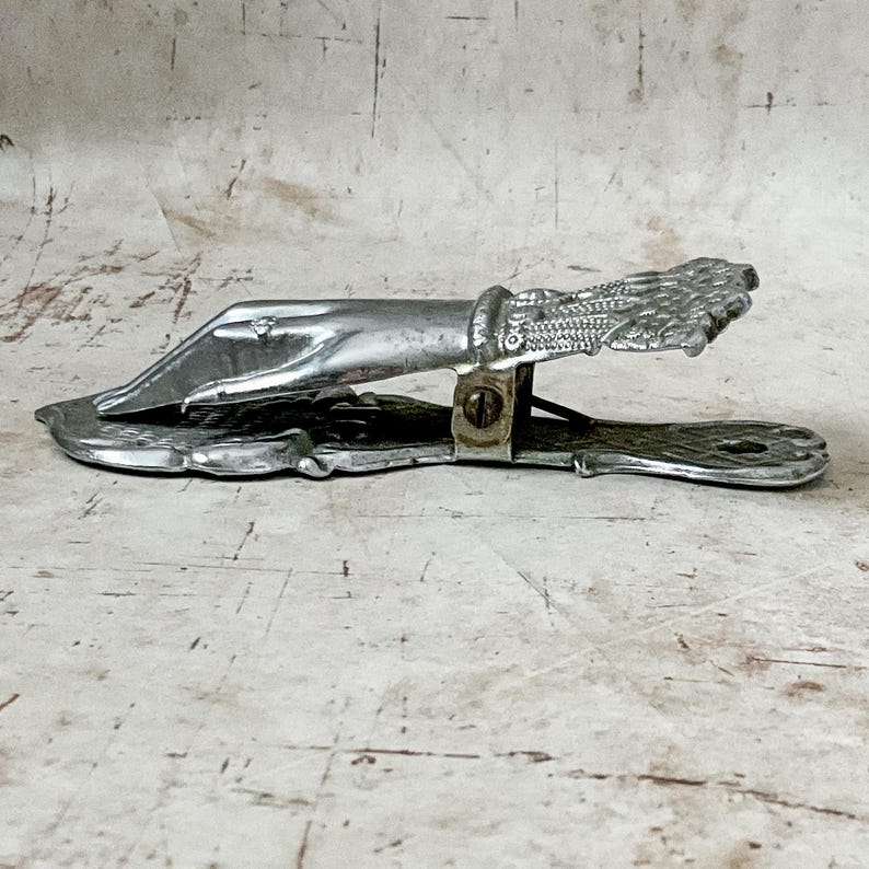 May include: A vintage silver-toned letter opener with a detailed hand and wing design. The opener features a blade and a clip mechanism, likely for holding papers. The design is ornate and the metal has a slightly aged appearance.