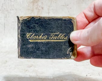 May include: A small, antique book with a dark blue cover and gold lettering that reads "Clarke's Tables." The book is held by a hand, showing its age and wear.