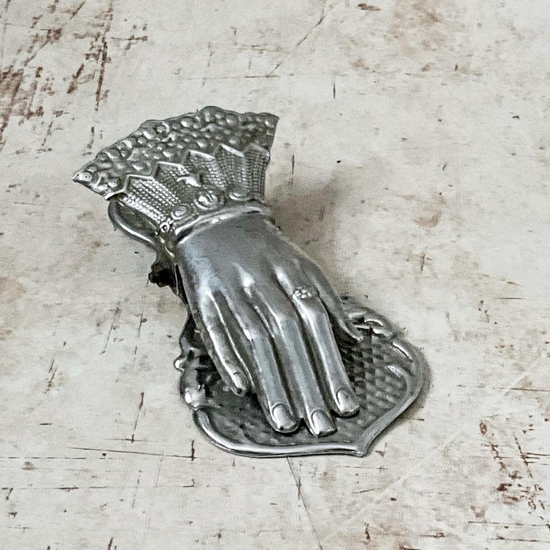 May include: A silver-toned hand-shaped clip, possibly a letter holder or decorative item. The hand is detailed with a ring and rests on a heart-shaped base. The clip has a decorative fan-like element at the wrist.