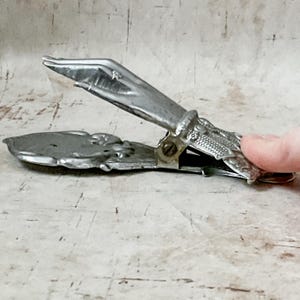 May include: Vintage silver-toned metal clip with ornate floral and textured details. The clip is open, revealing the interior mechanism. The design suggests it may be a paper clip or a similar office supply item.