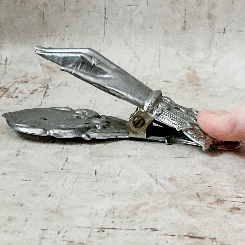 May include: Vintage silver-toned metal clip with ornate floral and textured details. The clip is open, revealing the interior mechanism. The design suggests it may be a paper clip or a similar office supply item.
