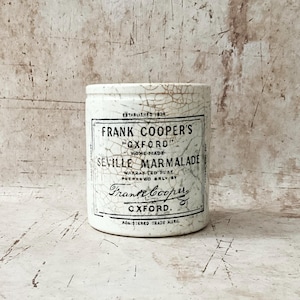 May include: A vintage white ceramic jar with a cracked glaze. The jar features a black and white label that reads "FRANK COOPER'S OXFORD SEVILLE MARMALADE" and "ESTABLISHED 1824". The jar is cylindrical and has a slightly rounded base.