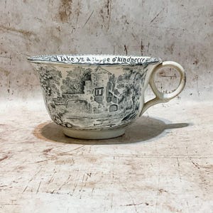 May include: A white and gray ceramic cup with a handle, featuring a black and white landscape scene with a castle and bridge. The rim of the cup has the text "take ye a cuppe o'kindnesse".