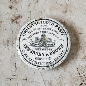 May include: A round, antique tin of "Oriental Tooth Paste" with black text on a white background. The text includes "Jewsbury & Brown, Chemists, 113 Market Street Manchester." A crest with lions and a crown is in the center.