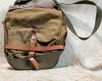 May include: An olive green canvas shoulder bag featuring a brown leather strap closure with metal rivets. The bag includes a wide, adjustable olive green webbing shoulder strap and a reinforced leather top trim with metal eyelets for secure fastening.